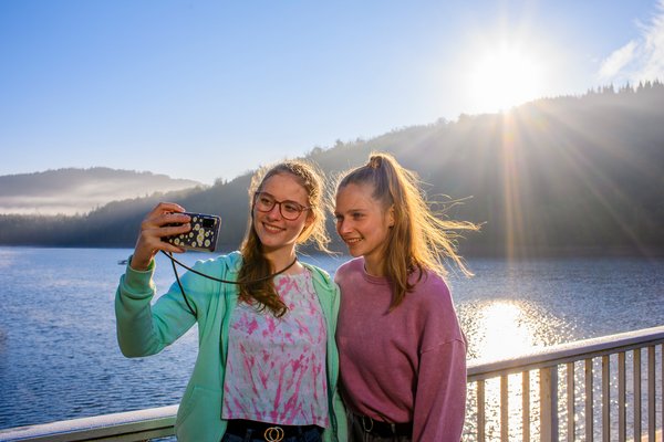 Zwei junge Frauen stehen auf dem Staudamm der Talsperre Nonnweiler. Im Hintergrund ist die Natur und das Wasser zu sehen. Sie selbst stehen im Vordergrund, eine hält ein Smartphone, mit dem sie ein Selfiebild machen.