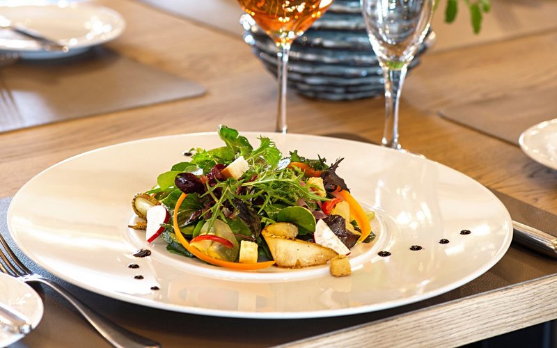 Auf dem Tisch im Restaurant des Victor's Seehotel Weingärtner stehen 2 gefüllte Weingläser. Im Vordergrund steht ein Teller mit einem Vorspeisensalat aus Rucula und Blattsalaten mit glasierten Birnen, Radieschen und Brotcroutons.