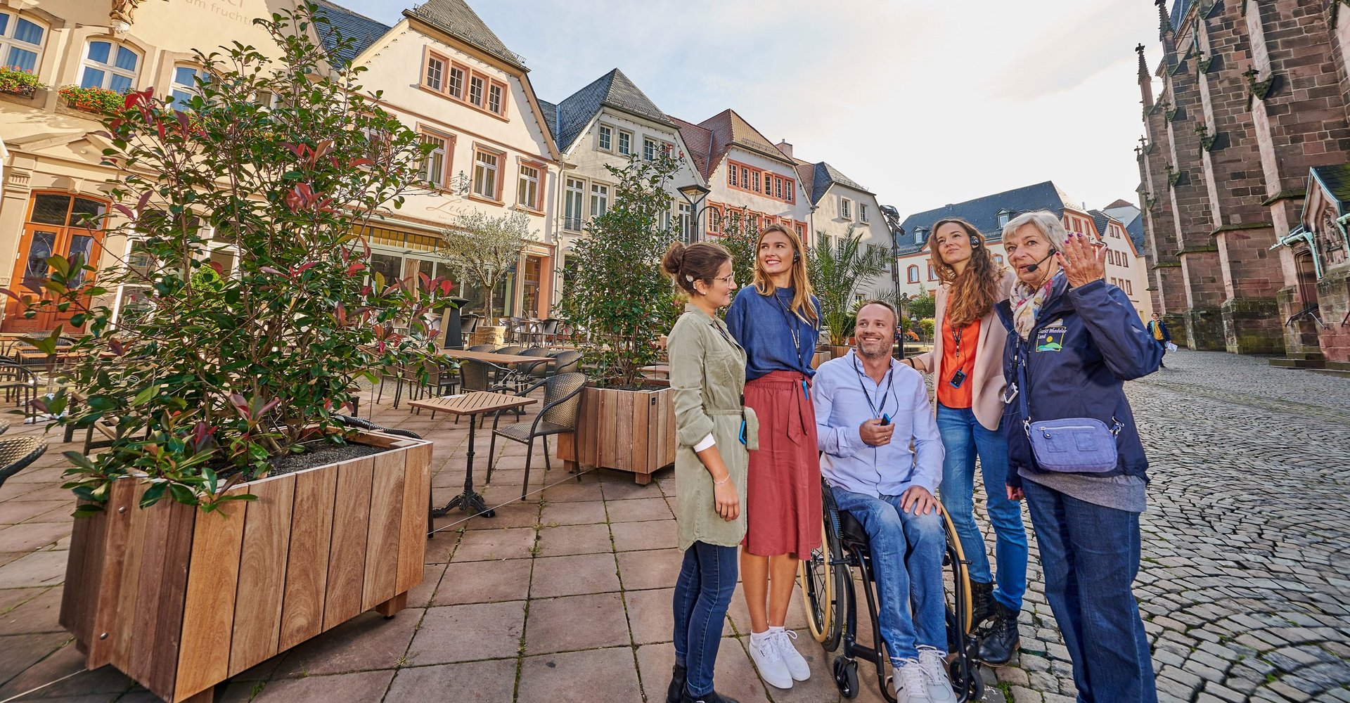 Eine Gästeführerin führt eine Gruppe durch die Stadt. Eine Person befindet sich im Rollstuhl.