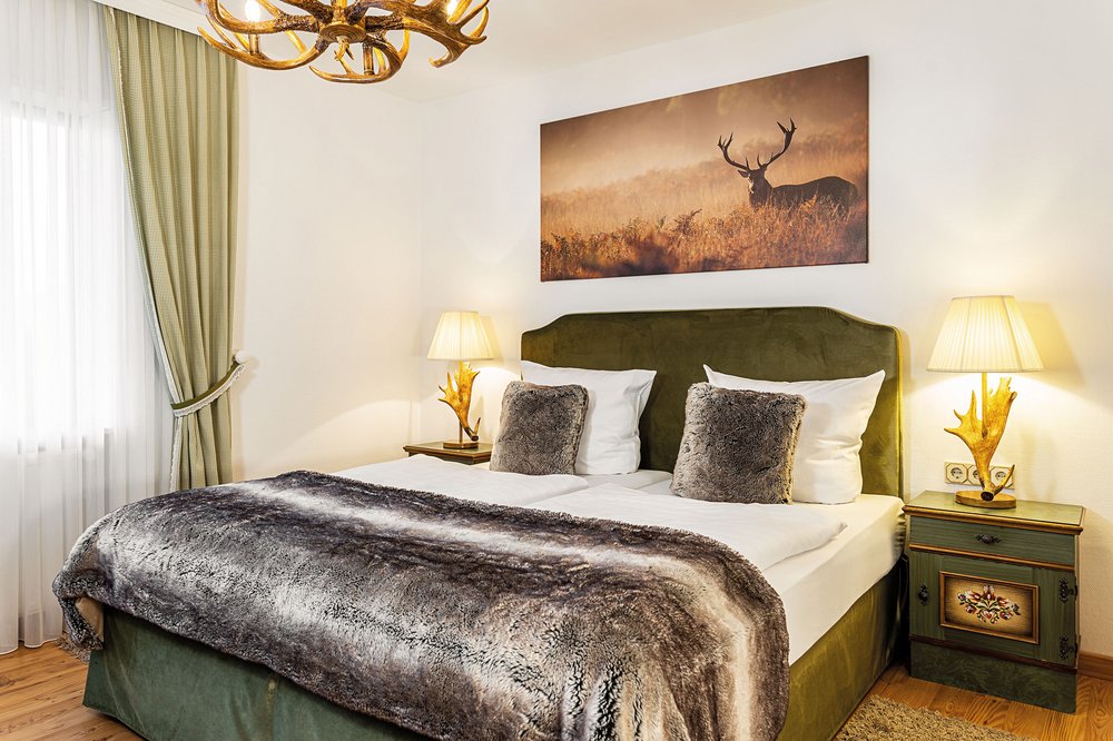 Landhausliebe im Victor's Seehotel Weingärtner Das Hotelzimmer des Victor's Seehotel Weingärtner ist im Landhausstil eingerichtet. Ein grünes Bett mit landhaustypischen Nachttischen und Lampen mit Hirschgeweih stehen im zentrum des Bildes. Alle Accessoires sind ebenfalls in Erdtönen und rustikal gehalten.