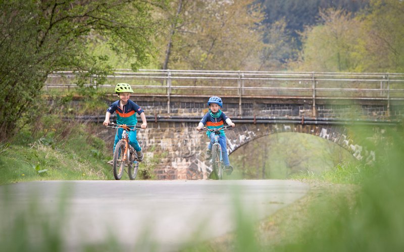 Zwei Kinder fahren mit Schwung auf ihren Fahrrädern über den asphaltierten Bahnradweg. 