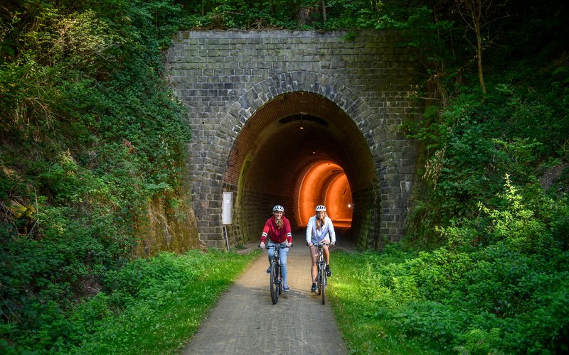 2 Radfahrer fahren auf dem Fritz-Wunderlich-Weg aus einem Tunnel aus Stein heraus. Der Tunnel ist im Inneren beleuchtet.