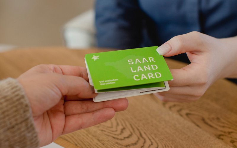 Eine Hand übergibt der anderen Hand ein grünes Kartenmäppchen der SaarlandCard.