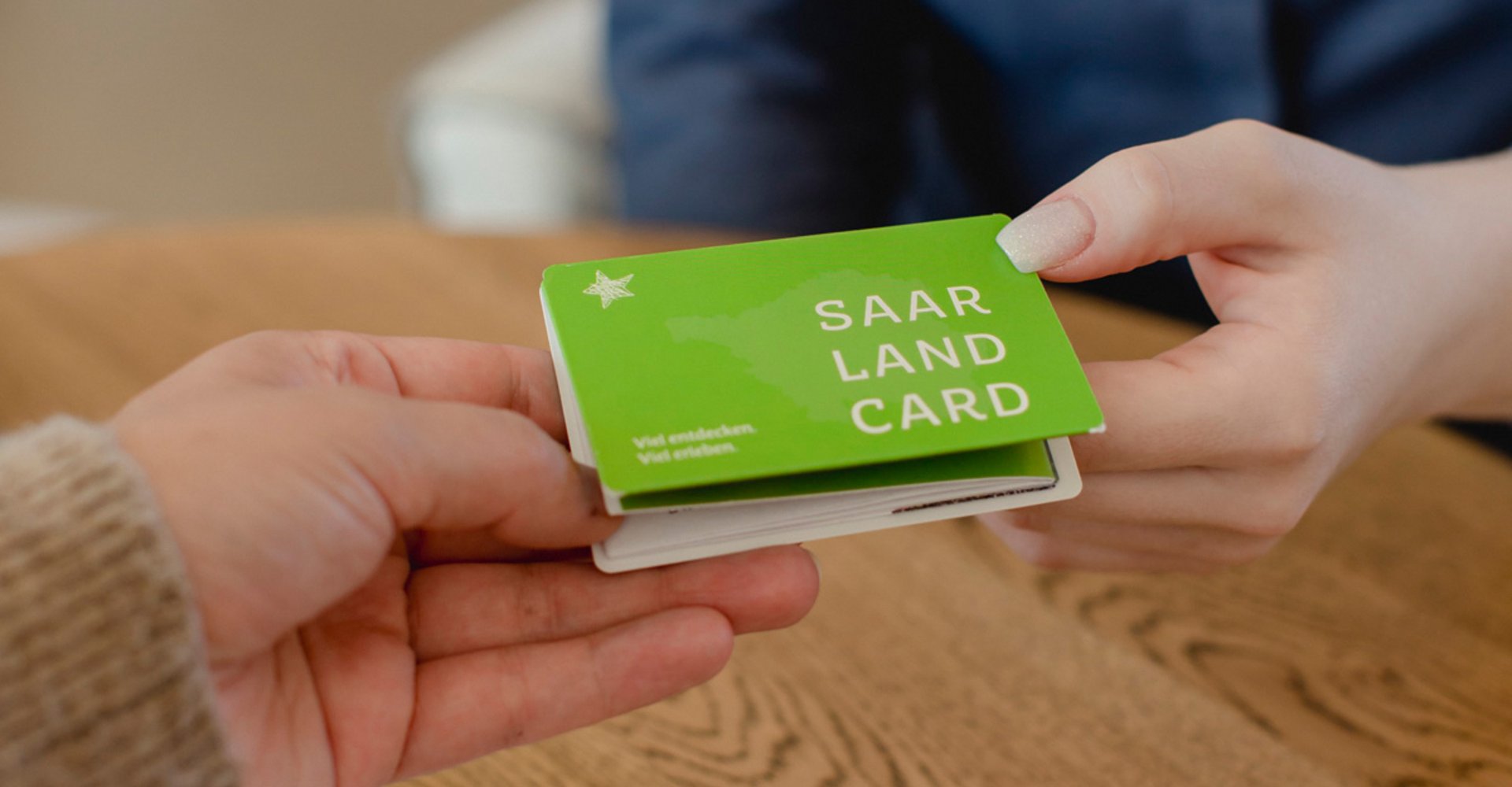 Eine Hand übergibt der anderen Hand ein grünes Kartenmäppchen der SaarlandCard.