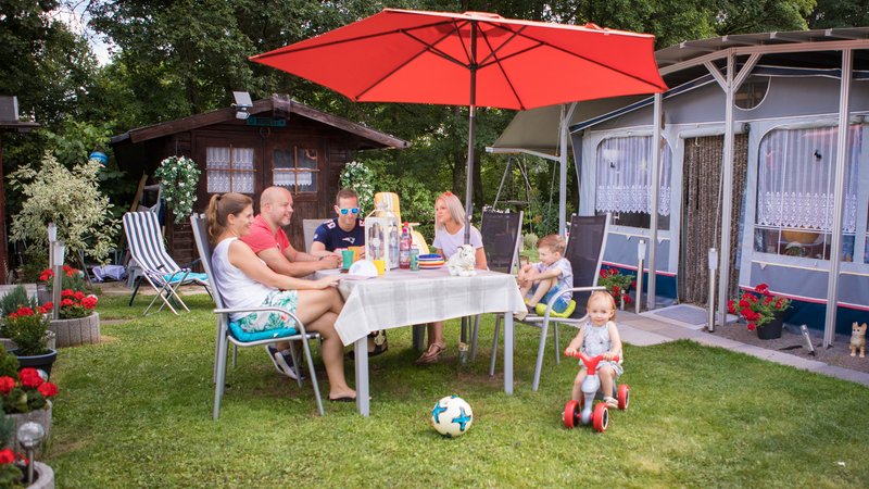 Zwei Paare sitzen vor einem Wohnwagen mit Vorzelt sowie einem Gartenhaus aus Holz an einem Gartentisch mit Sonnenschirm und unterhalten sich. Zwei Kinder sind am Spielen.