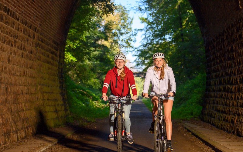 Zwei Radfahrer fahren auf dem Fritz-Wunderlich-Weg in einen steinernen, beleuchteten Tunnel hinein.