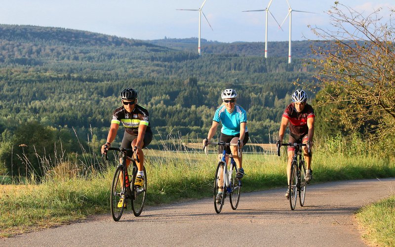 3 Rennradfahrer, zwei Männer und eine Frau, fahren eine asphaltierte Straße entlang und genießen die schöne Aussicht.