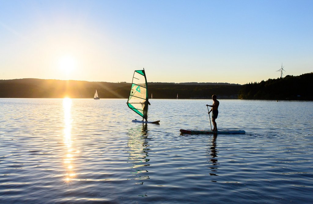 © Klaus-Peter Kappest Ein Windsurfer und ein Stand-Up-Paddler gleiten bei Sonnenuntergang gemächlich über den Bostalsee.