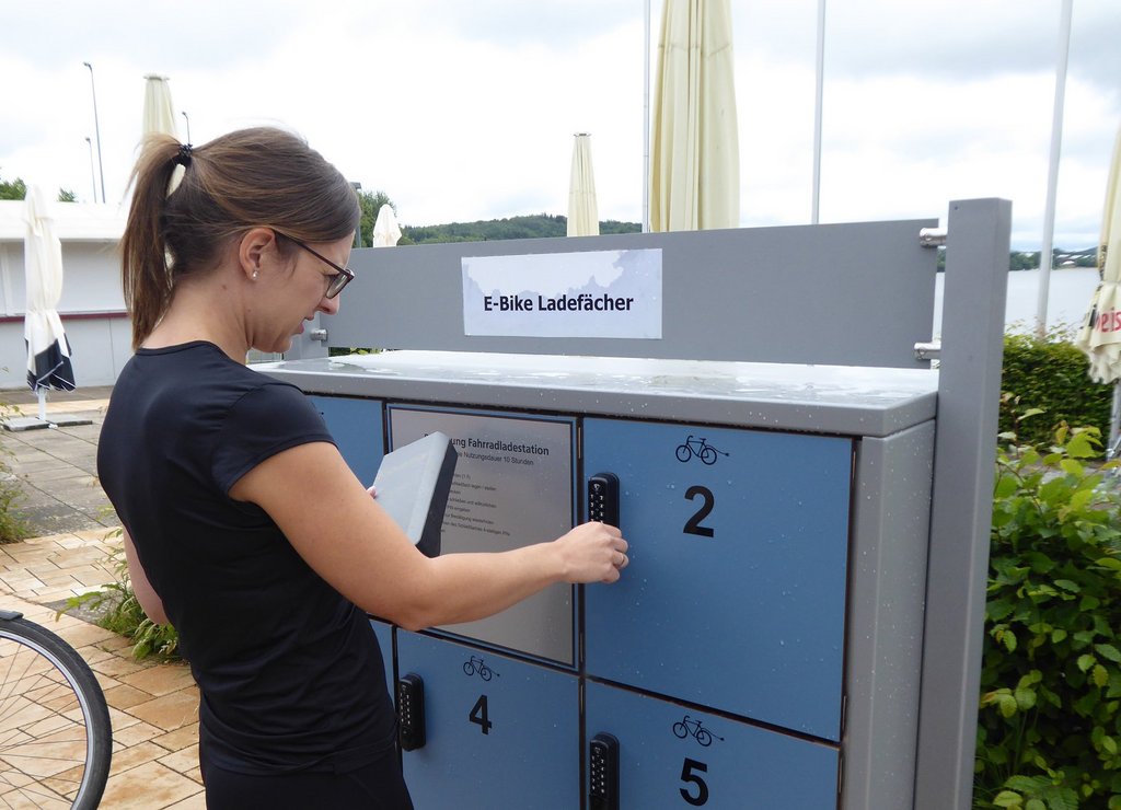 E-Bike-Ladestationen am Bostalsee Eine Frau mit Pferdeschwanz und Brille ist gerade dabei, ein Fach der E-Bike-Ladestation am Bostalsee zu öffnen.