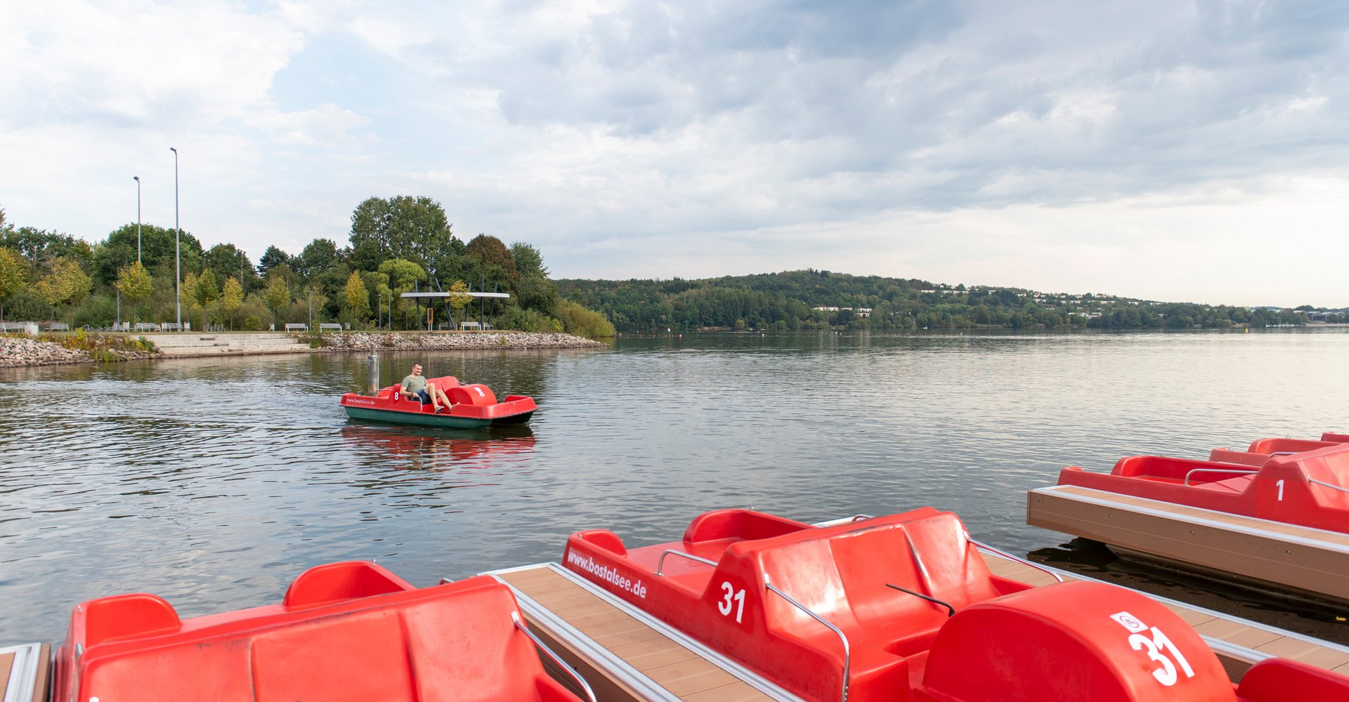 Neben der Anlegestelle mit den roten Tretbooten gleitet ein weiteres Tretboot mit einem Mann über das Wasser.