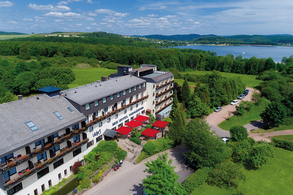 Eingebettet in die grüne Naturlandschaft des Bostalsees steht das Victor's Seehotel Weingärtner. Die Aufnahme zeigt, das im Landhausstil gebaute Gebäude aus der Vogelperspektive. Im Hintergund ist ein Teil des Bostalsees zu sehen.