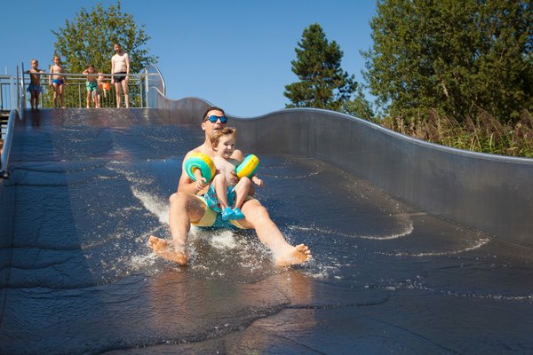 Badespaß auf der Wellenrutsche im Strandbad Bosen Ein junger Mann rutscht mit Kleinkind in Schwimmflügeln eine wellenförmige Wasserrtusche herunter. Oben, am Rand der Rutsche warten weitere Personen, um ebenfalls herunterrutschen zu können.