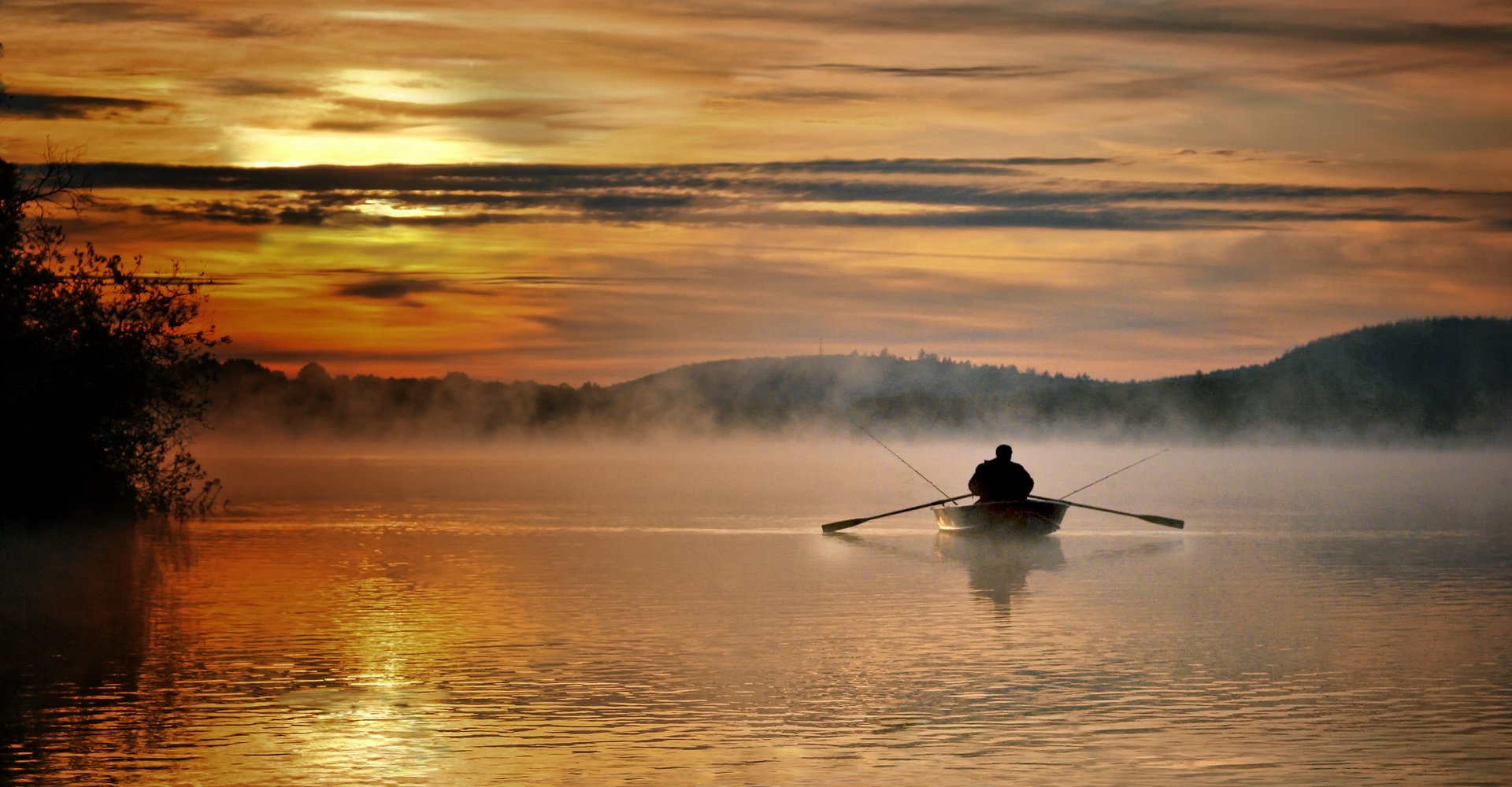 Ein Angler rudert mit seinem Boot über den Bostalsee. Das Bild ist durch die Einwirkung der Sonne komplett in Orange getaucht.