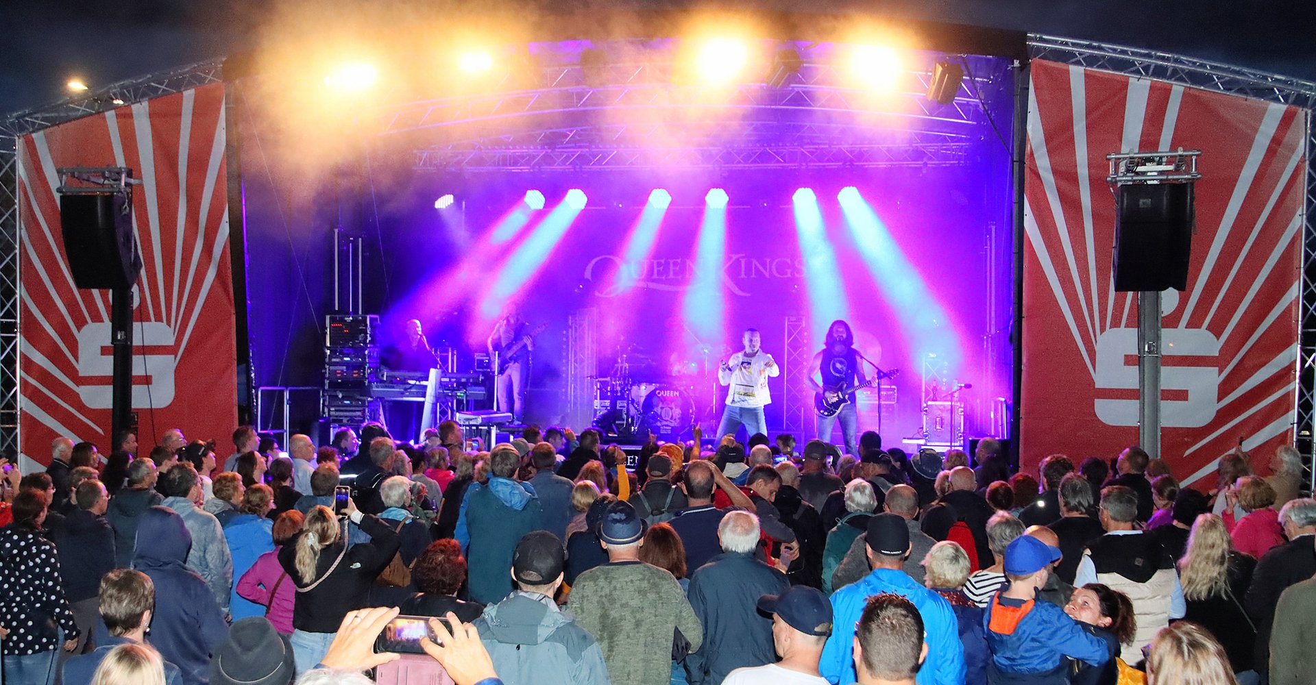 Viele Menschen stehen vor einer Bühne, werden vom Bühnenlicht angestrahlt und bewegen sich zur Musik der Band, die auf der Bühne spielt.