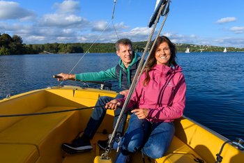 Ein Paar mittleren Alters sitzt am Heck eines gelben Segelboots. Die Dame hält die Seile der Segel, während der Herr das Segelboot am Heckruder steuert. Im Hinergrund sind der Bostalsee mit weiteren Segelbooten sowie das Seeufer zu sehen.